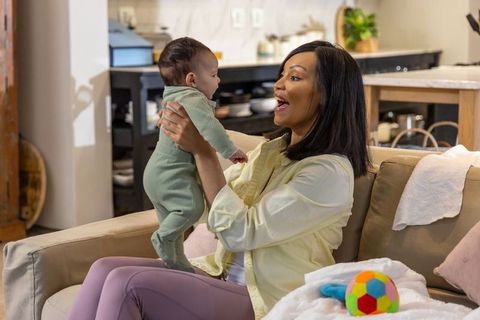 African american mother holding smiling infant girl on sofa in cozy open-plan living room
