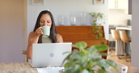 Woman working from home sipping coffee at laptop on dining table in open-plan interior