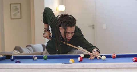 Man with dreadlocks leaning over blue-felt pool table aiming precise shot in home lounge