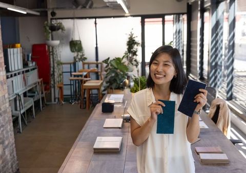 Smiling asian designer presenting teal and navy fabric swatches in bright textile studio