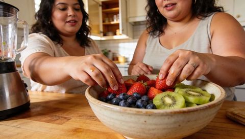 Preparing colorful fresh fruit bowl together on wooden kitchen island with blender, jars