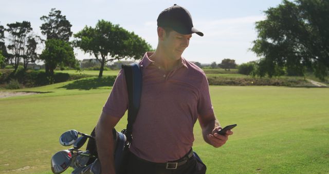 Male golfer carrying golf bag, checking smartphone, standing on green golf course on sunny day. Ideal for topics about technology in sports, leisure activities, or promoting golf equipment and accessories.