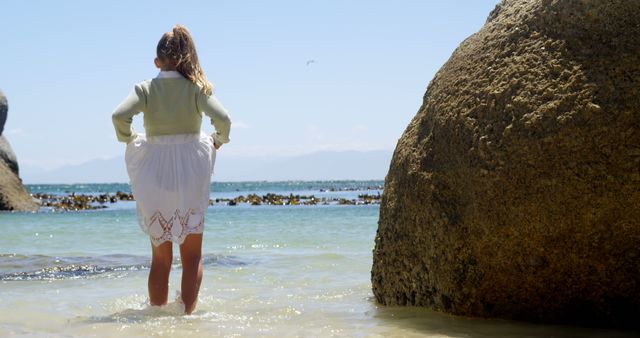 Back view of caucasian girl in white skirt standing in water next to big stone. Childhood, free time, summer, travel, vacations and lifestyle, unaltered.