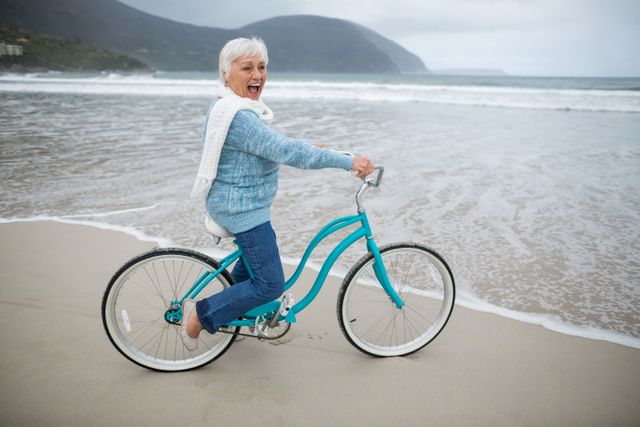 Senior woman enjoying a ride on a bicycle along the beach, smiling and looking joyful. Ideal for promoting active lifestyles, retirement communities, healthy living, and leisure activities for seniors. Can be used in advertisements, brochures, and websites focusing on senior wellness and outdoor activities.