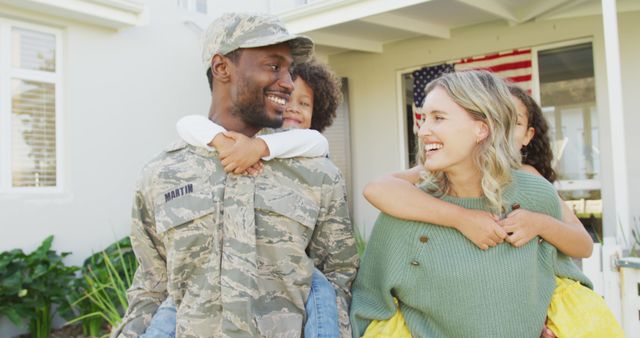 Image portrays a joyful reunion of a mixed race family. The military father wears a camouflage uniform and holds one child on his back while the other rejoins the family. The American flag is visible in the background, signifying patriotism. Ideal for use in topics related to military homecoming, family bonding, patriotism, and support for military families.