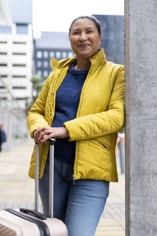 Mature woman in yellow jacket leaning on post holding luggage at urban transit plaza