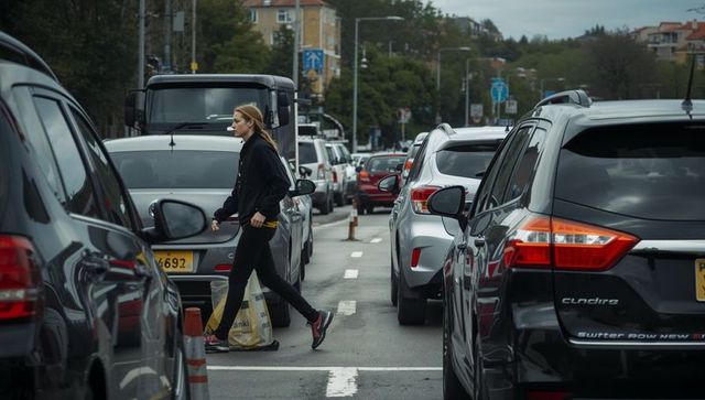 Woman crossing busy city street between stopped cars during rainy traffic jam carrying shopping bag