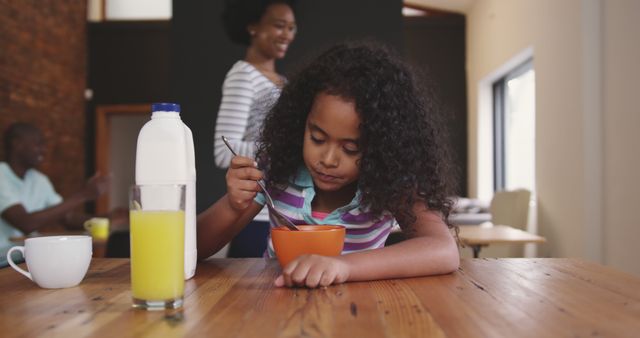 Young girl enjoying breakfast at home, with family in background. Milk, orange juice and cereal on table. Perfect for family, home life, breakfast time advertising, parenting blogs, and nutritional content.