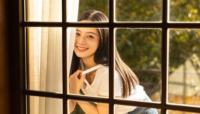 Young woman smiling through window grid during golden hour, cozy home lifestyle
