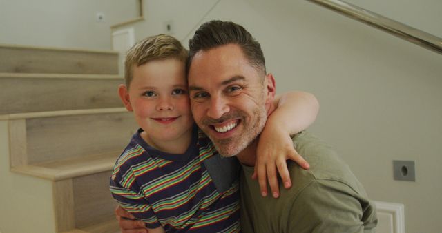 This image shows a joyful moment between a father and his son hugging on a staircase at home. Both are smiling warmly, indicating a close bond and family joy. Ideal for use in advertisements, parenting articles, family blogs, and social media posts highlighting familial love and bonding.