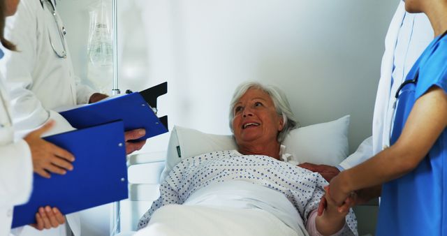 Elderly woman lying in hospital bed smiling while talking to medical staff holding clipboards. Perfect for topics related to healthcare, patient recovery, nursing care, elderly care, doctor-patient relationship, hospital settings, and medical consultations.