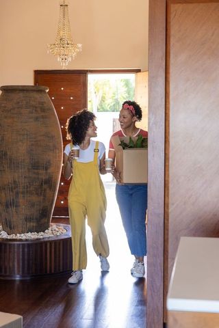 Diverse friends moving in carrying box with plant and coffee in sunlit entryway