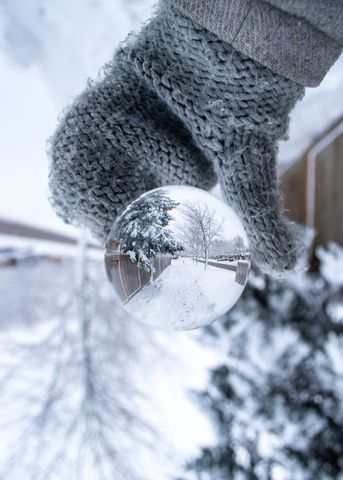 Wool glove holding glass sphere reflecting winter sidewalk with snowy trees and fence