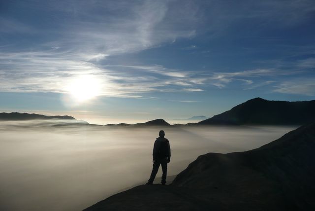 Silhouetted hiker stands on mountain ridge at dawn, overlooking fog-covered landscape with rising sun. Ideal for use in travel blogs, outdoor adventure promotions, motivational posters, and nature-inspired advertisements.