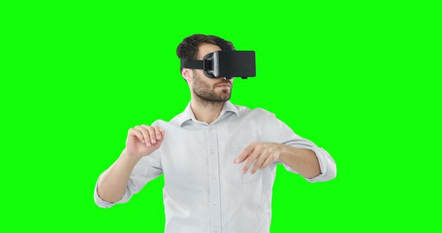 This image shows a man in a white shirt using a virtual reality headset against a green screen background. His hands are raised, as though interacting with a virtual environment. Suitable for themes related to technology, virtual reality, innovation, and gaming. Can be used for educational materials, presentations, blogs, and advertisements highlighting virtual reality experiences.