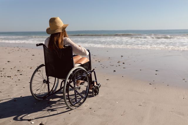 Disabled woman wearing a hat sitting in a wheelchair on a sandy beach, looking at the ocean. Ideal for use in articles or advertisements promoting accessibility, travel, independence, and outdoor activities for people with disabilities. Can also be used in lifestyle blogs, travel websites, and health and wellness campaigns.