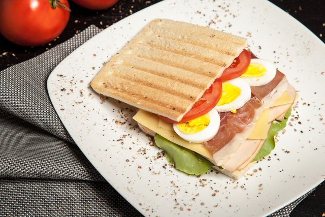 This high-quality image showcases a perfectly grilled panini sandwich placed on a white plate. The sandwich is filled with sliced boiled eggs, juicy tomatoes, and rich cheese, topped with crispy lettuce. Generously seasoned with black pepper, this panini looks both delicious and wholesome. Ideal for illustrating recipe blogs, restaurant menus, and nutrition articles focusing on healthy meals and gourmet snacks.