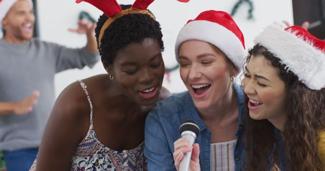 Diverse group of friends singing into a microphone at a holiday party. They are wearing Santa and reindeer hats, appearing joyful and festive. The background includes holiday decorations. Ideal for promoting holiday events, social gatherings, or festive season celebrations.