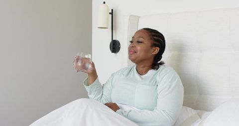 Mid adult african american woman relaxing in bed holding glass of water in mint pajamas