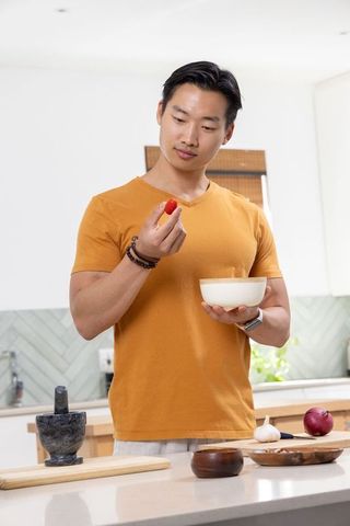 Asian man inspecting cherry tomato while holding bowl in modern kitchen