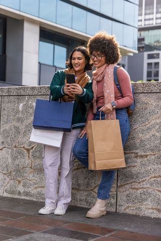 Two women leaning on stone wall checking smartphone with shopping bags in urban plaza