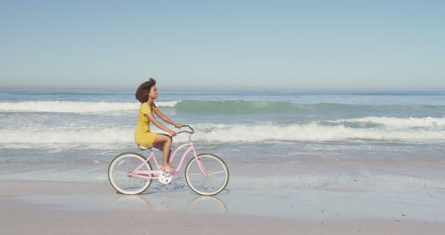 Happy biracial woman riding bicycle on sunny beach by the sea. Summer, free time, relaxation and vacations.