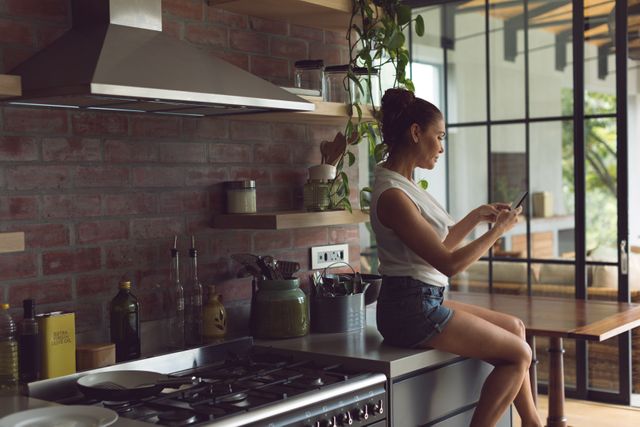 Side view of beautiful woman using mobile phone while sitting on worktop in kitchen at comfortable home