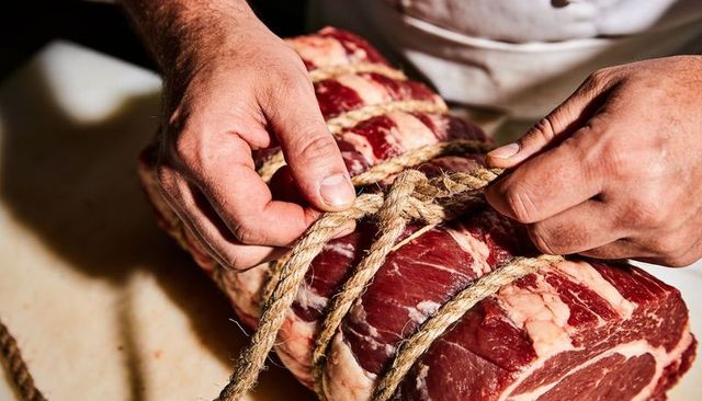 Butcher tying twine around rolled beef roast on cutting board, hands knotting
