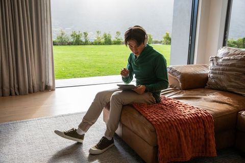 Asian man using tablet with stylus on leather sofa in modern home office with lawn view
