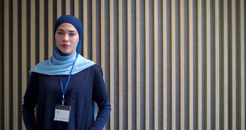 Confident muslim woman wearing hijab and conference badge standing by vertical wood panels