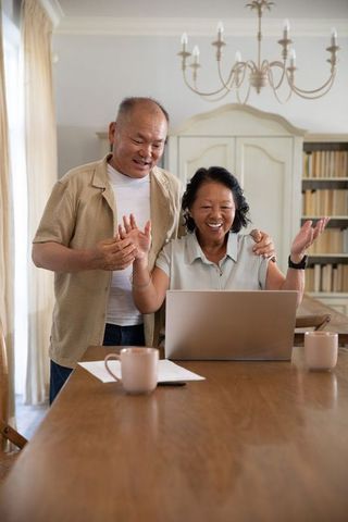 Senior asian couple celebrating video call using laptop at cozy dining table