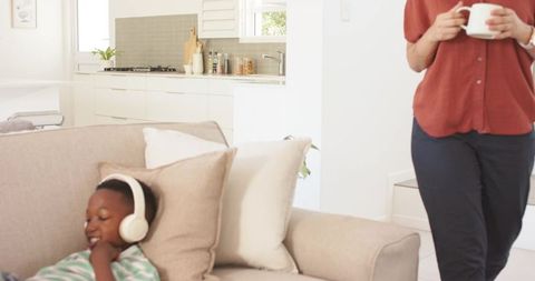 Relaxing teen wearing headphones on sofa in sunlit open-plan living room, adult holding mug