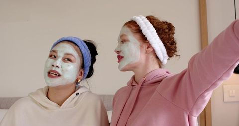 Asian and white friends wearing face masks and headbands taking selfie in cozy loungewear