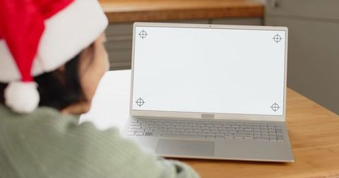 Senior asian woman in santa hat sitting at kitchen table facing laptop with blank screen