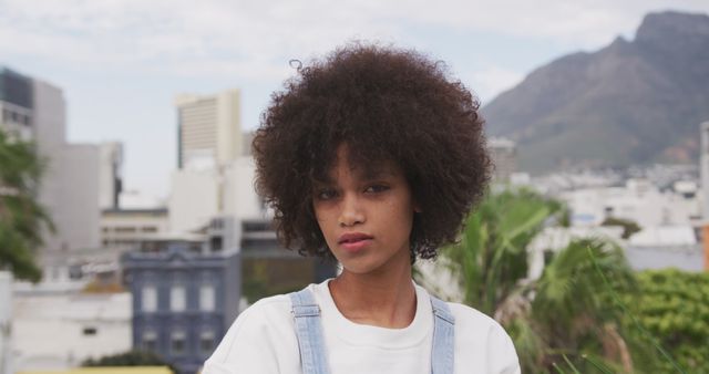 Young woman with afro hair standing confidently in an outdoor urban area. She is wearing casual clothing and appears self-assured. The background features city buildings, trees, and mountains, making it ideal for use in advertisements, lifestyle articles, fashion blogs, and social media posts that focus on urban life, personal style, or natural beauty.