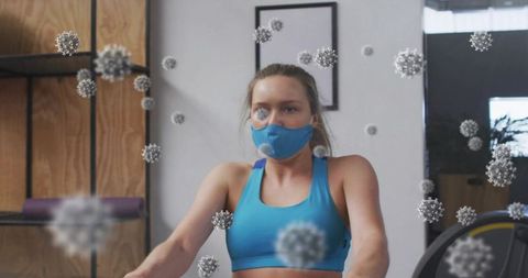 Woman wearing mask on rowing machine surrounded by virus particles, indoor gym workout