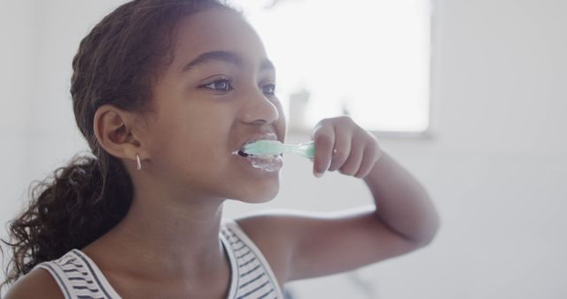 Young girl engaging in daily hygiene routine by brushing her teeth in the bathroom. Ideal for use in advertisements and articles focused on children’s health, dental hygiene, parenting tips for morning routines, and personal care products.