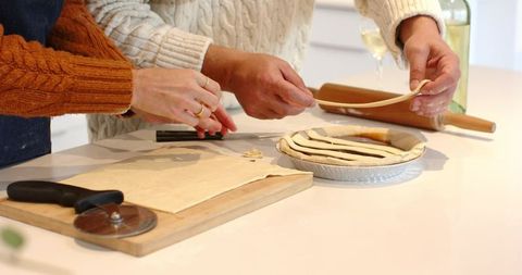 Couple assembling lattice-top pie wearing knit sweaters on bright kitchen counter