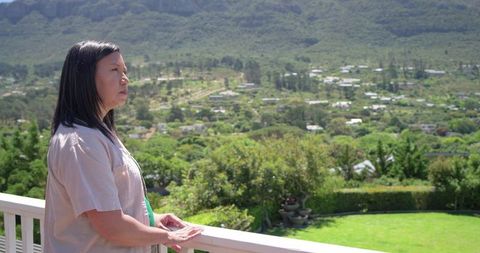 Asian woman standing on balcony resting hands on railing gazing over lush valley