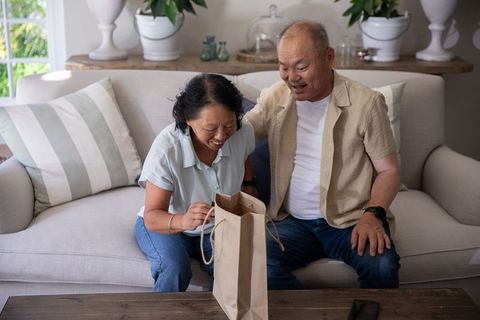 Senior asian couple opening gift bag and sharing joyful moment on cozy living room sofa