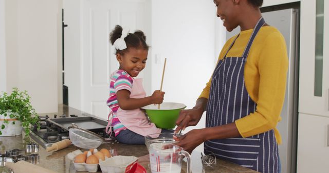 This warm and joyful image showcases an African American mother and daughter enjoying quality time together while cooking in their home kitchen. They are both wearing aprons, suggesting they are invested in their culinary activity. This image is perfect for use in family-oriented content, cooking and recipe blogs, advertisements focusing on family bonding or parental involvement, and any material aiming to depict the warmth and love shared within family relationships, especially during times of isolation or lockdown.