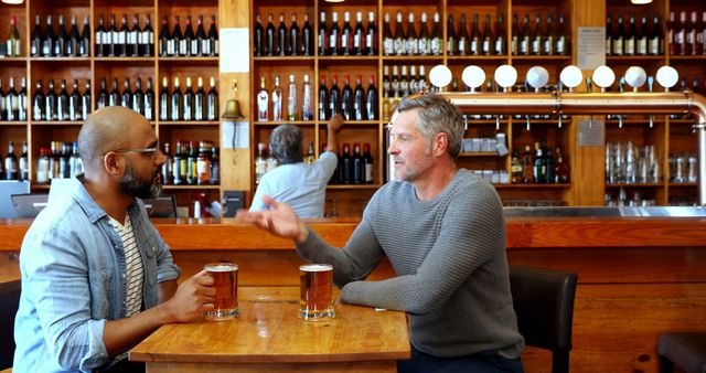 Two friends sitting at a wooden table in a cozy pub, enjoying a beer and engaging in conversation. Perfect for themes of friendship, socializing, relaxation, leisure time, and pub environments. Useful for articles or advertisements related to breweries, pubs, or social gatherings.