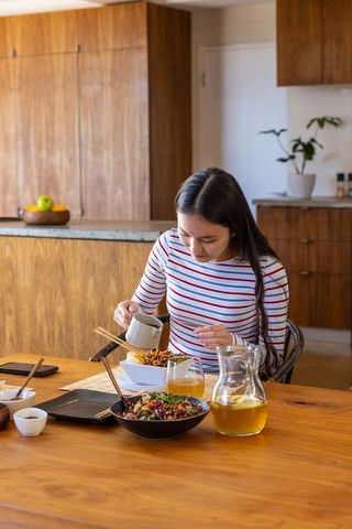 Woman adding sauce to asian noodle bowl in cozy home dining area