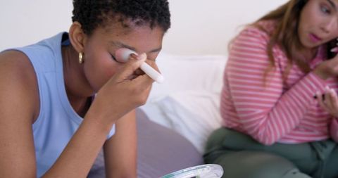 African american friends applying eye makeup on bed sharing compact mirror and brushes