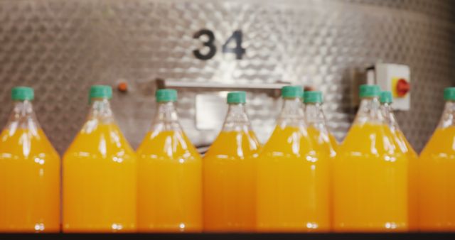 This image depicts multiple bottles of orange juice lined up, freshly filled in a juice production facility. They are arranged uniformly, indicating a large-scale manufacturing process. The bottles have green caps and the background showcases industrial equipment, hinting at the location being a factory or a juice processing plant. This image can be useful for articles, presentations, or advertisements related to the food and beverage industry, manufacturing processes, or healthy lifestyle products.