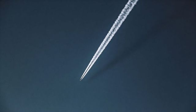 High-altitude jet cutting diagonal path across deep blue sky leaving twin contrails and shadow