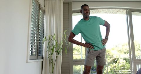Mid-adult African American man stretching at home by sliding doors with balcony view