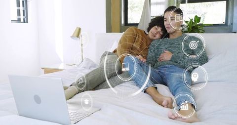 Relaxed same-sex couple reclining on bed using laptop with futuristic hud technology overlay
