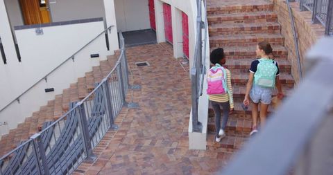 Two diverse female friends walking up brick school steps with backpacks