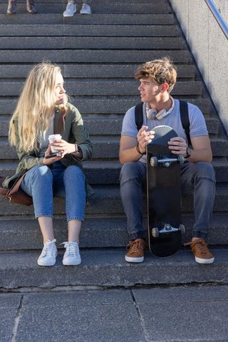 Teenage friends sitting on city steps holding skateboard and coffee, casual conversation